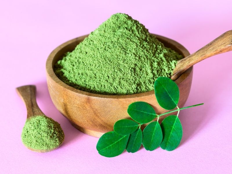 Moringa powder in a wooden bowl with fresh moringa leaves on a pink background – natural breast milk increasing powder for lactating mothers
